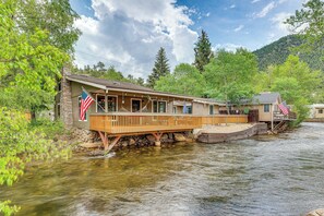 Eine Flusskabine mit Holzterrasse und amerikanischen Flaggen, umgeben von üppigem Grünland und einem Bergpanorama.
