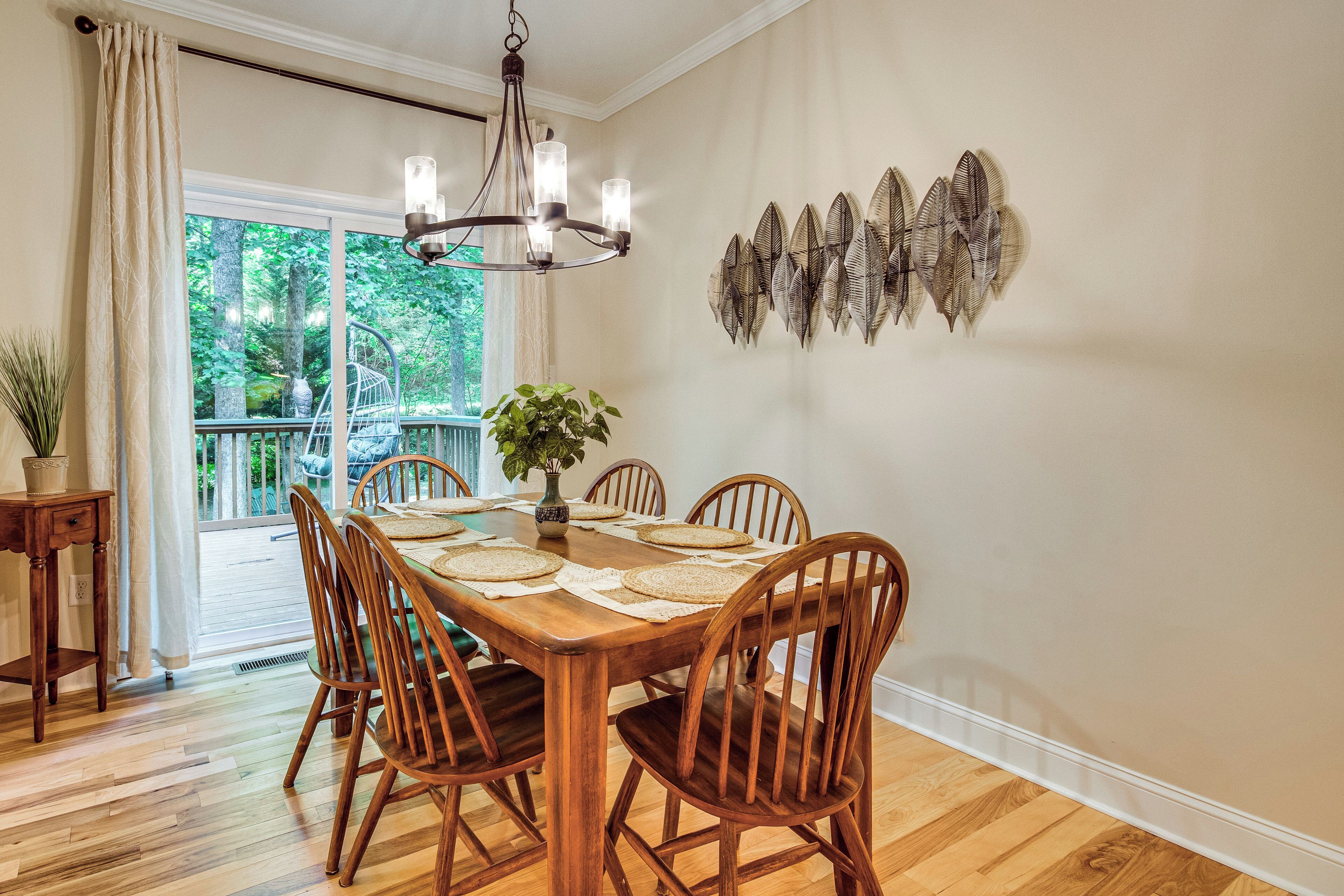 A dining room with a wooden table set for six, surrounded by wooden chairs, a chandelier, and a decorative wall piece.