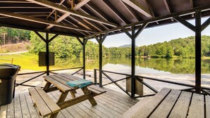 Une terrasse en bois couverte, dotée de tables de pique-nique, qui offre une vue sur un lac paisible et des collines boisées.
