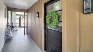 A hallway with a wooden door, a green wreath, and a view into a room with a window and a chair. - 1 Block to Beach & Eats: Modern Indialantic Apt (Indialantic)