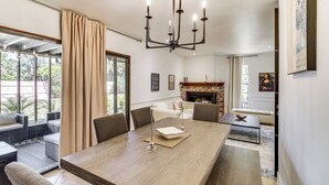 A dining area with a large wooden table, chairs, and a chandelier. There is a fireplace in the background and a view of the outdoors through large windows. - 1 Block to Beach & Eats: Modern Indialantic Apt (Indialantic)