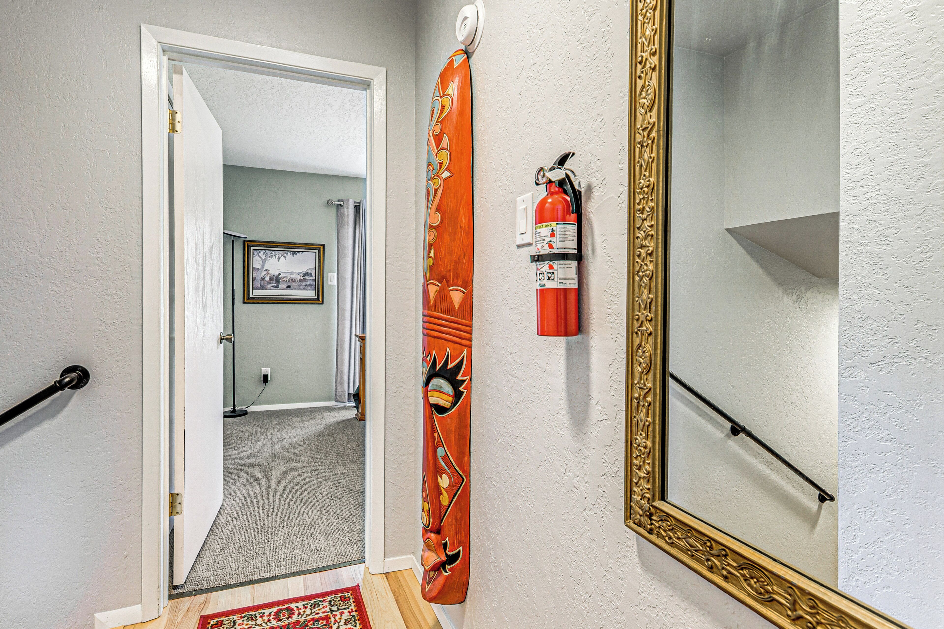 A hallway with a wooden door, a fire extinguisher, a mirror, and a framed picture on the wall.