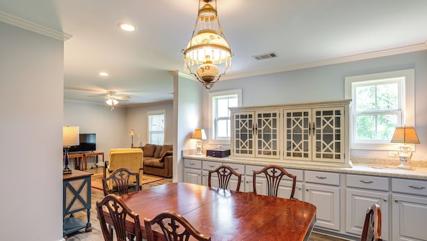 A dining area with a wooden table and chairs, a chandelier, and a kitchen with white cabinets and a window. - 7 Mi to Ole Miss: Quiet Rural Retreat w/ Yard! (Oxford)
