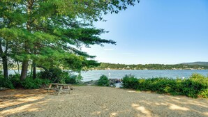 A sandy beach with a picnic table and trees, overlooking a body of water and a distant shoreline. - Lake & Beach Access: Family Townhome in Laconia (Laconia)