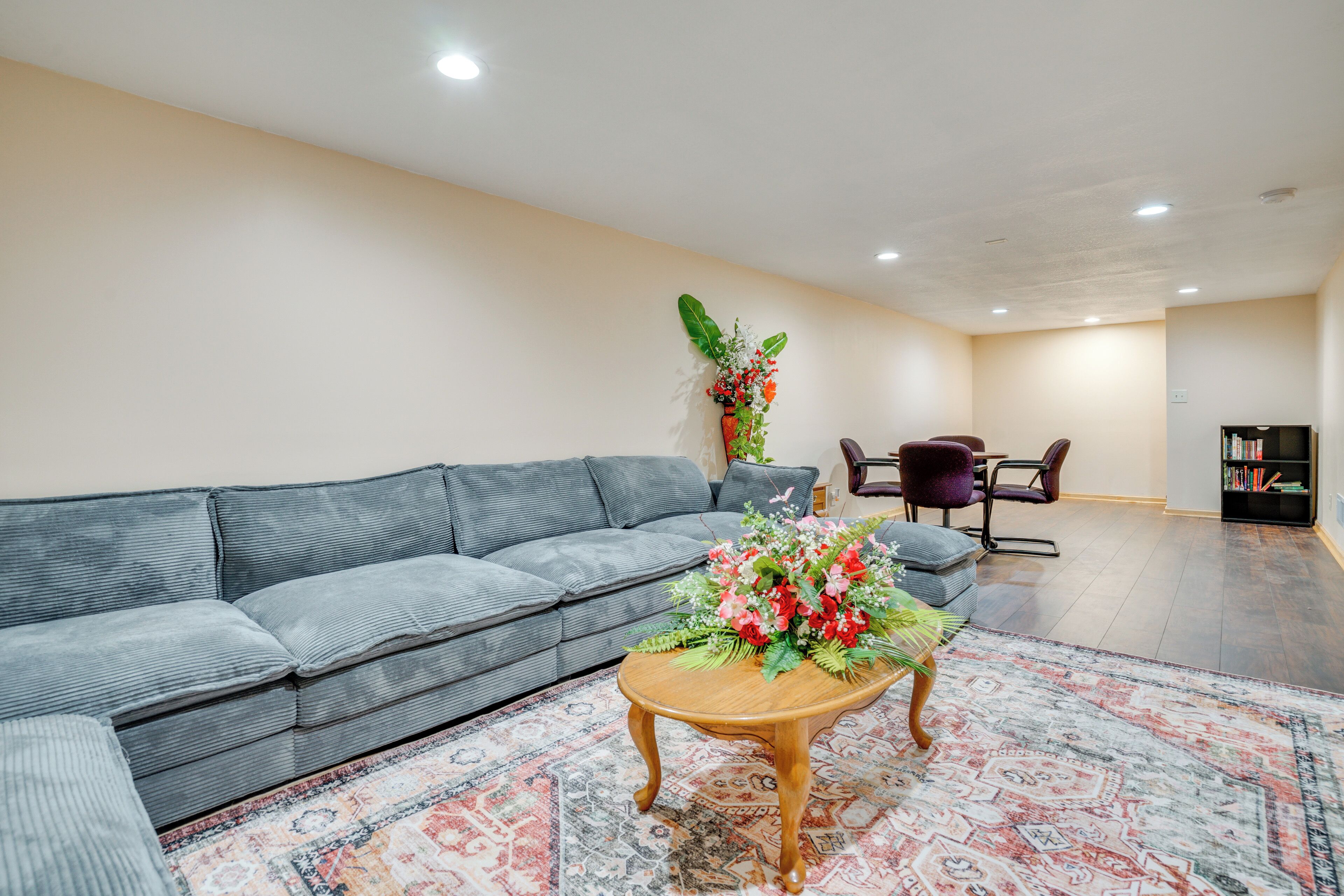 Uma sala de estar moderna com um sofá cinzento, uma mesa de centro de madeira com um arranjo floral, um tapete estampado e uma estante de livros ao fundo.