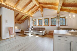 A spacious room with a high wooden ceiling, a large window with a view of trees, a sofa, and a kitchen area.