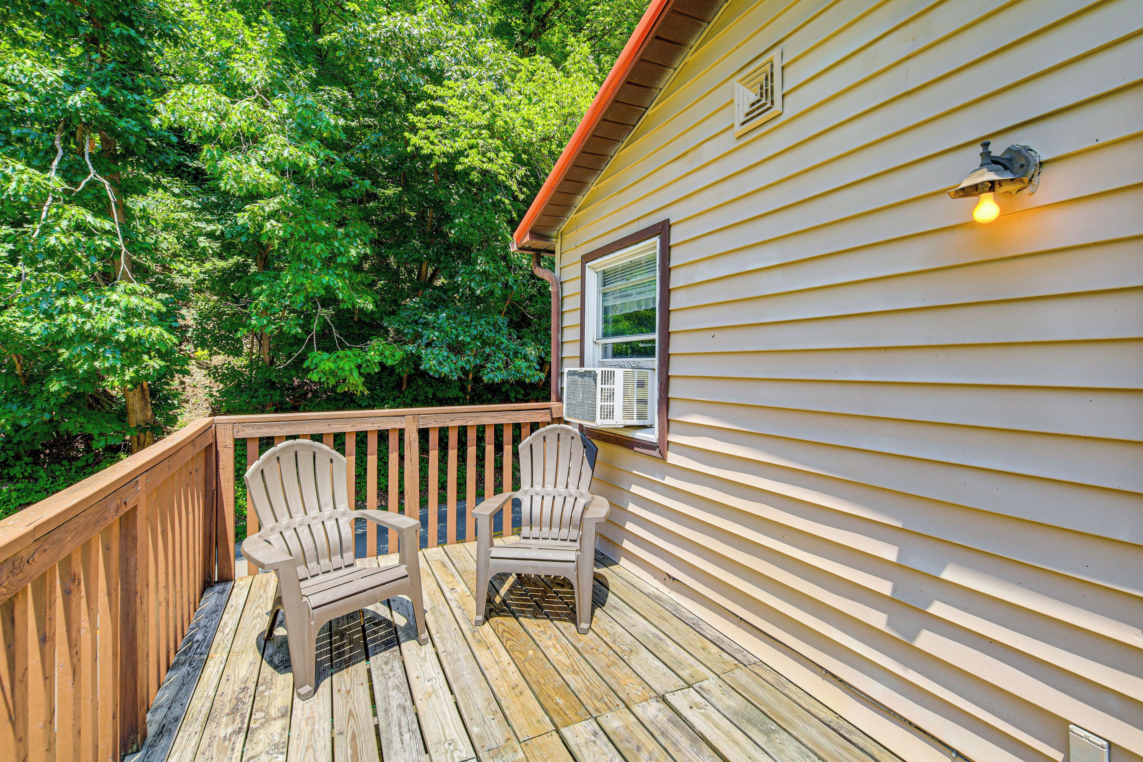 A wooden deck with two Adirondack chairs, a window with an air conditioning unit, and a light fixture on the side of a house surrounded by trees.