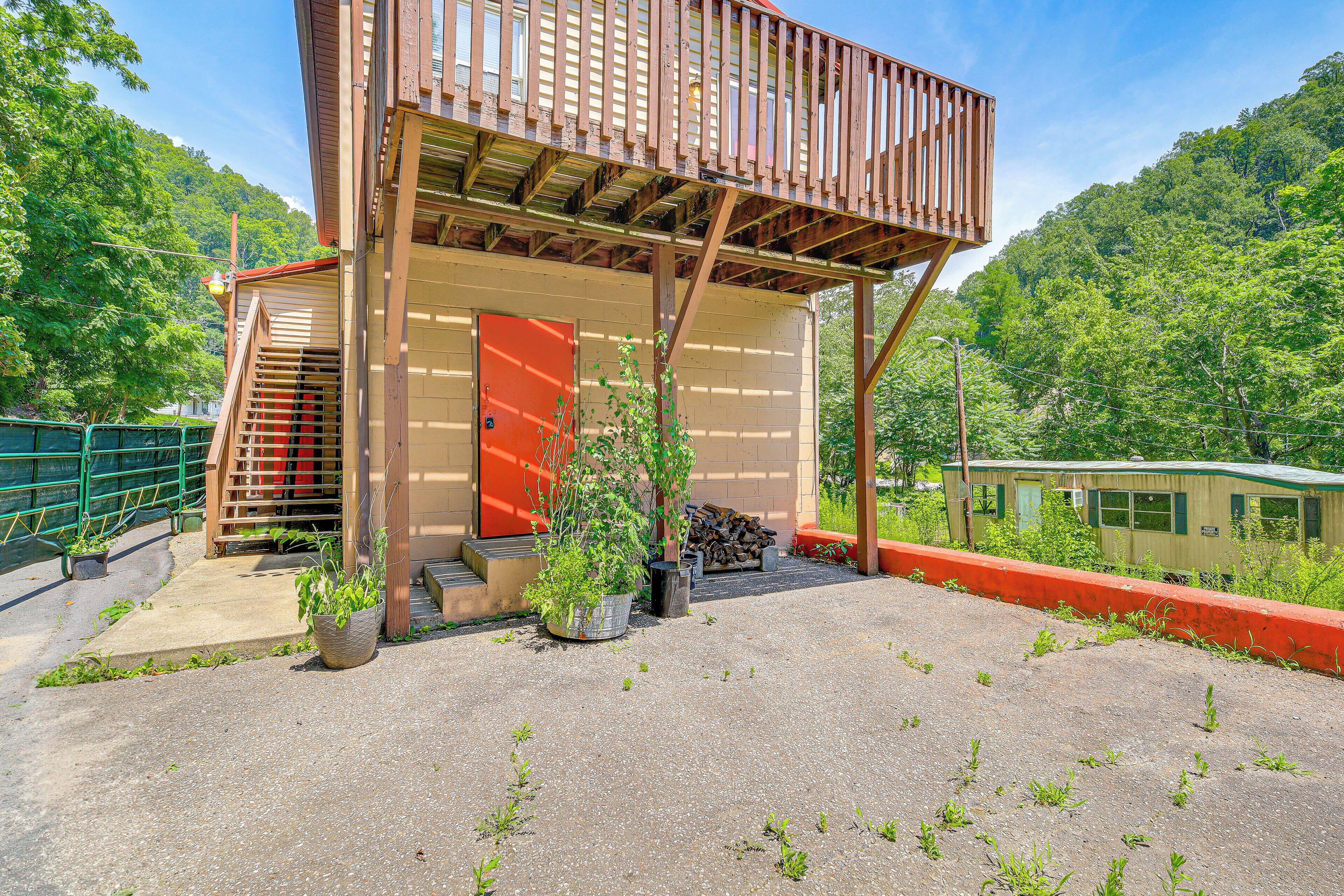 Une structure en bois avec une porte rouge et un balcon, entourée de verdure, avec un train visible au loin.