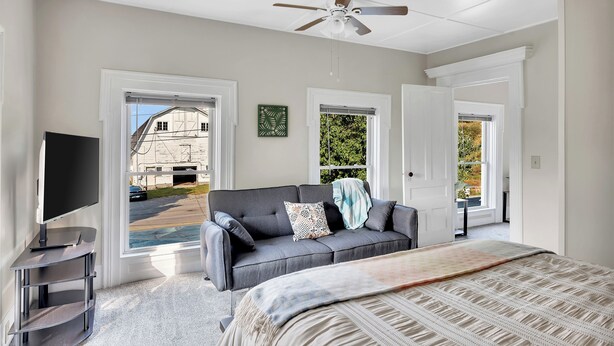 A bedroom with a grey sofa, a bed with a patterned bedspread, a ceiling fan, and large windows with views of a building and greenery. - Renovated 1800s Brick Farmhouse in Granville (Granville)