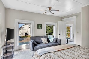A bedroom with a grey sofa, a bed with a patterned bedspread, a ceiling fan, and large windows with views of a building and greenery. - Renovated 1800s Brick Farmhouse in Granville (Granville)
