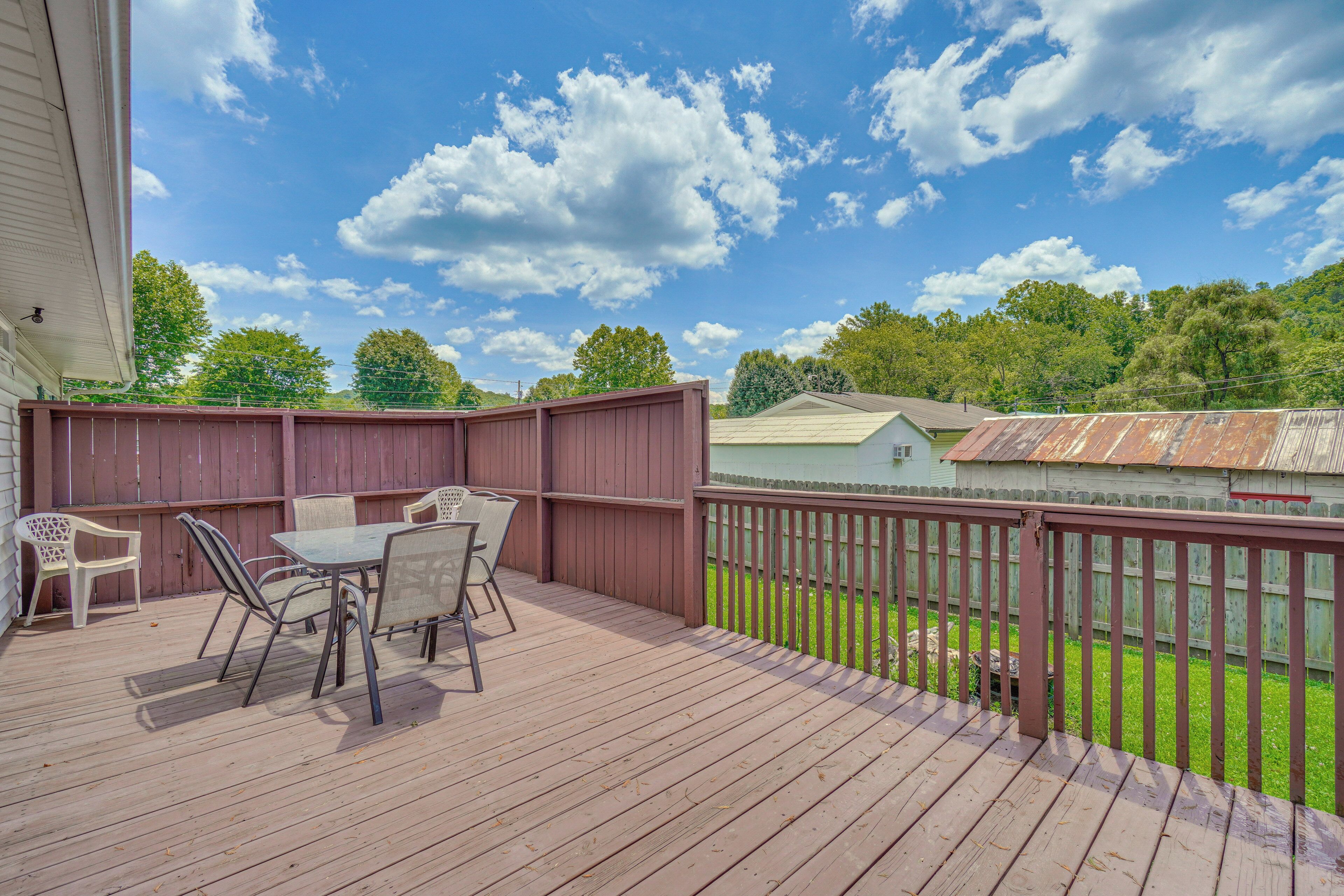 A wooden deck with a table and chairs, surrounded by a wooden fence, overlooking a grassy yard and a house with a metal roof.