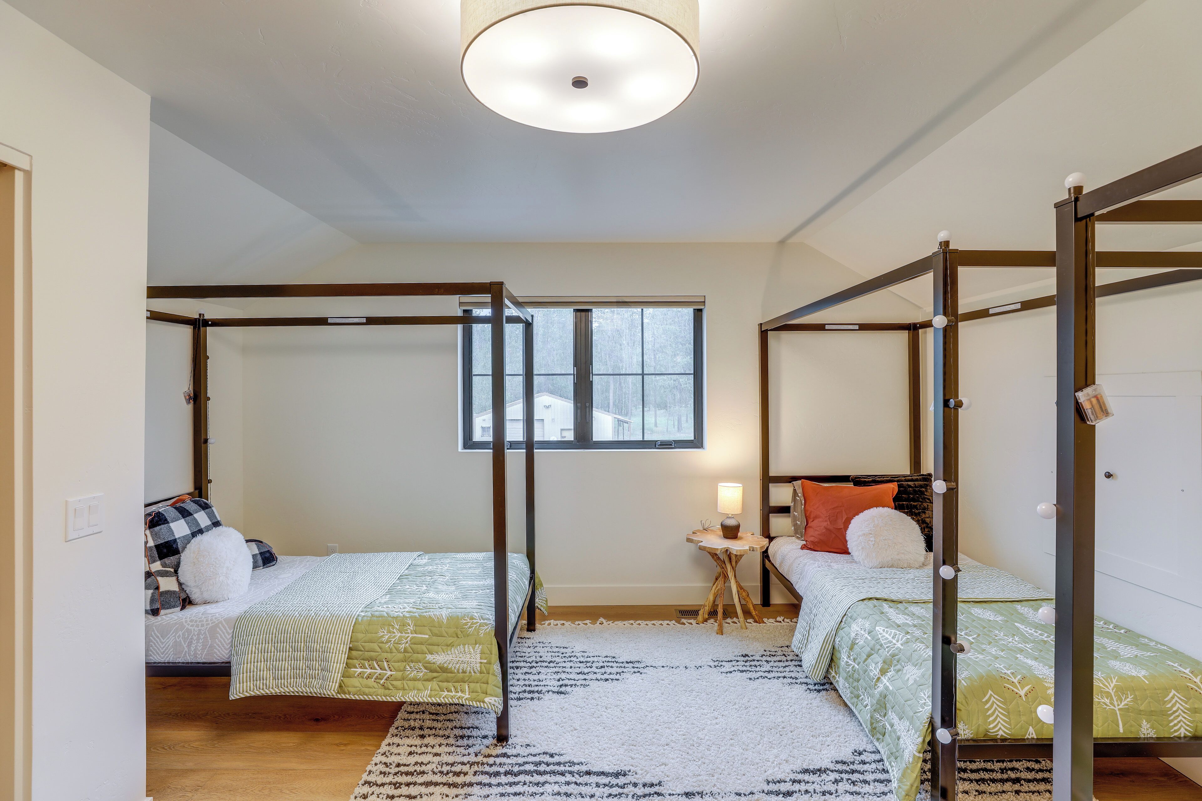 A room with two beds, a window, a small table with a lamp, and a ceiling light.