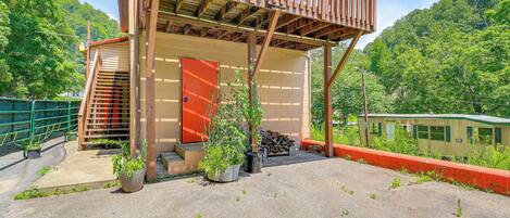 Une structure en bois avec une porte rouge et un balcon, entourée de verdure, avec un train visible au loin.