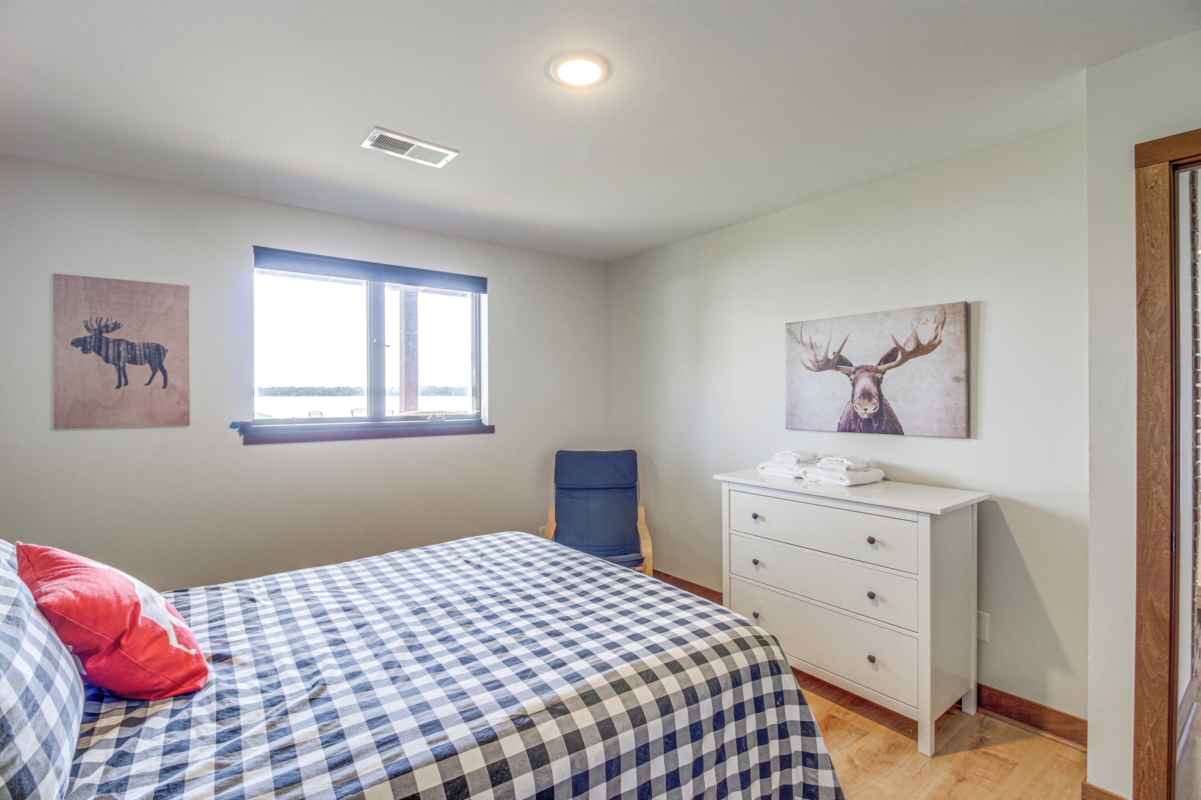 A bedroom with a checkered bedspread, a window with blinds, a desk with a telephone, and two wall-mounted animal paintings.