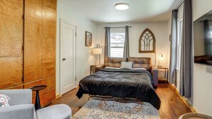 Ein Schlafzimmer mit großem Kleiderschrank, einem Bett mit dunkler Bettdecke, einem Nachttisch mit Lampe und einem Fenster mit Vorhängen.