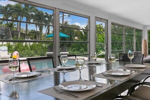 A dining area with a long table set for four, large windows offering a view of palm trees and greenery, and a poolside area with a pink flamingo decoration.