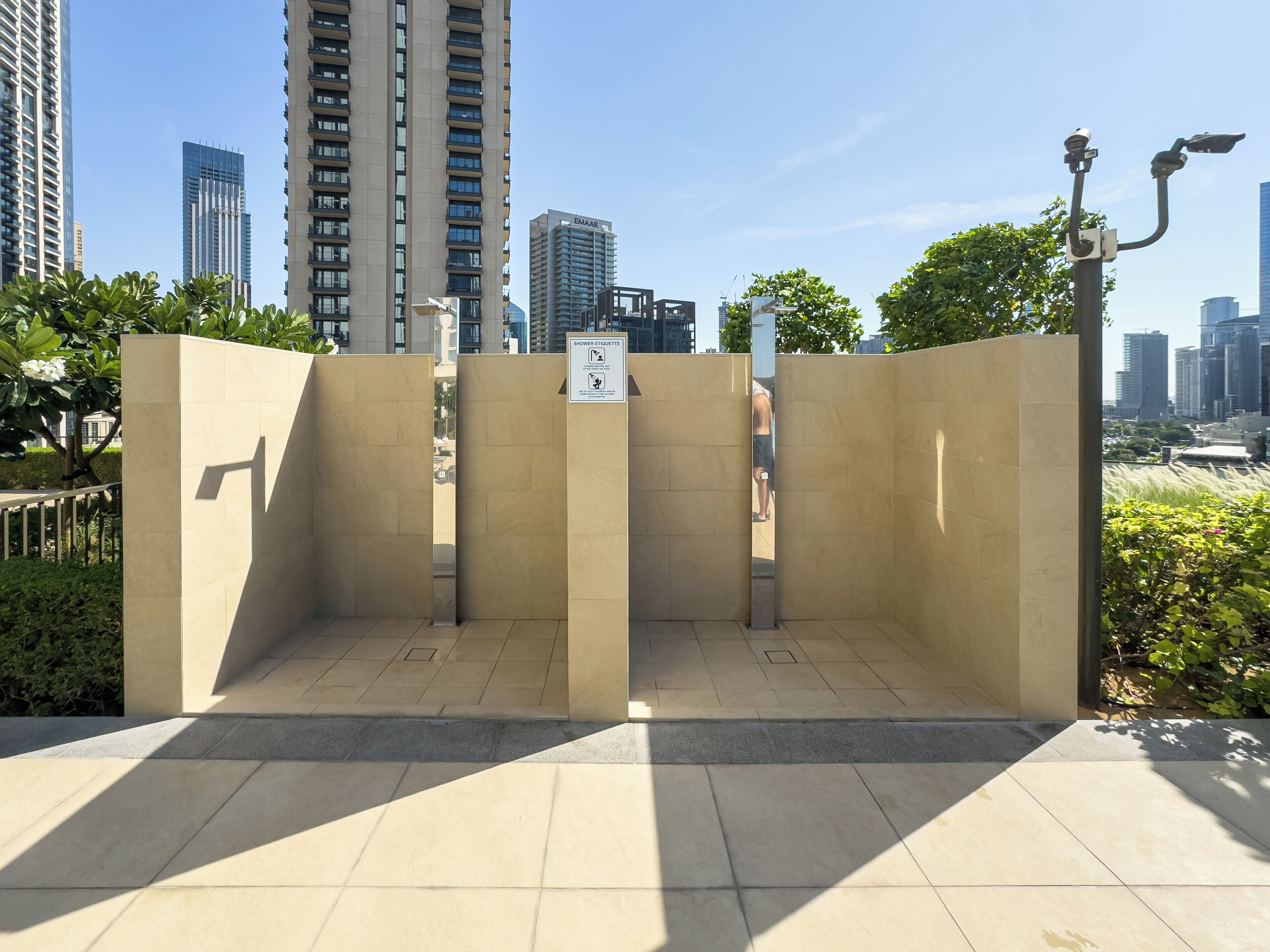 A public restroom with a 'Shower Etiquette' sign, surrounded by a tiled floor and a metal fence, with tall buildings in the background.