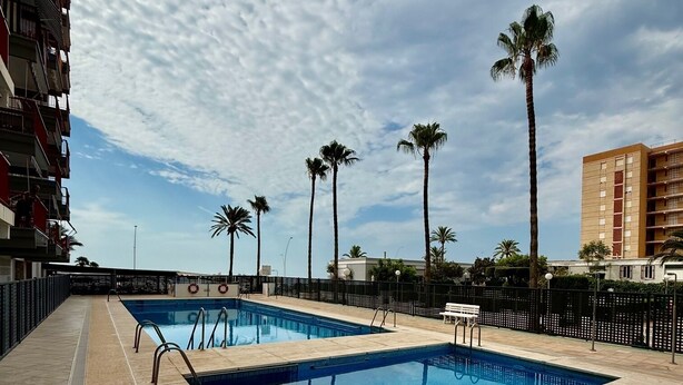 A swimming pool area with two pools, palm trees, and a building in the background. - Apartamento Torreblanca III Ref 063 (Torreblanca)
