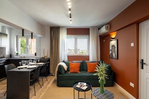 A modern living room with a green sofa, dining table, and chairs. There is a small table with a book and a plant. The room has a large window with curtains, an air conditioning unit, and a framed picture on the wall. - Colorful Bright 2BR on Iconic Calea Victoriei (Bucharest)