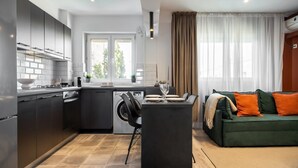 Eine moderne Küche mit grauen Schränken, einem eingebauten Herd und einem Essbereich mit Tisch und Stühlen. Im Wohnbereich befindet sich ein grünes Sofa mit orangefarbenen Kissen.