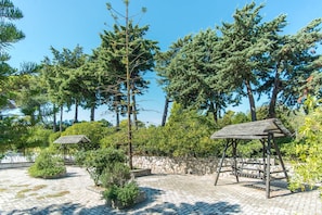 A paved outdoor area with stone walls and wooden gazebos, surrounded by tall trees and greenery.