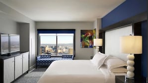A hotel room with a large bed, a television, a window with a city view, a blue patterned bench, and a colorful abstract painting.
