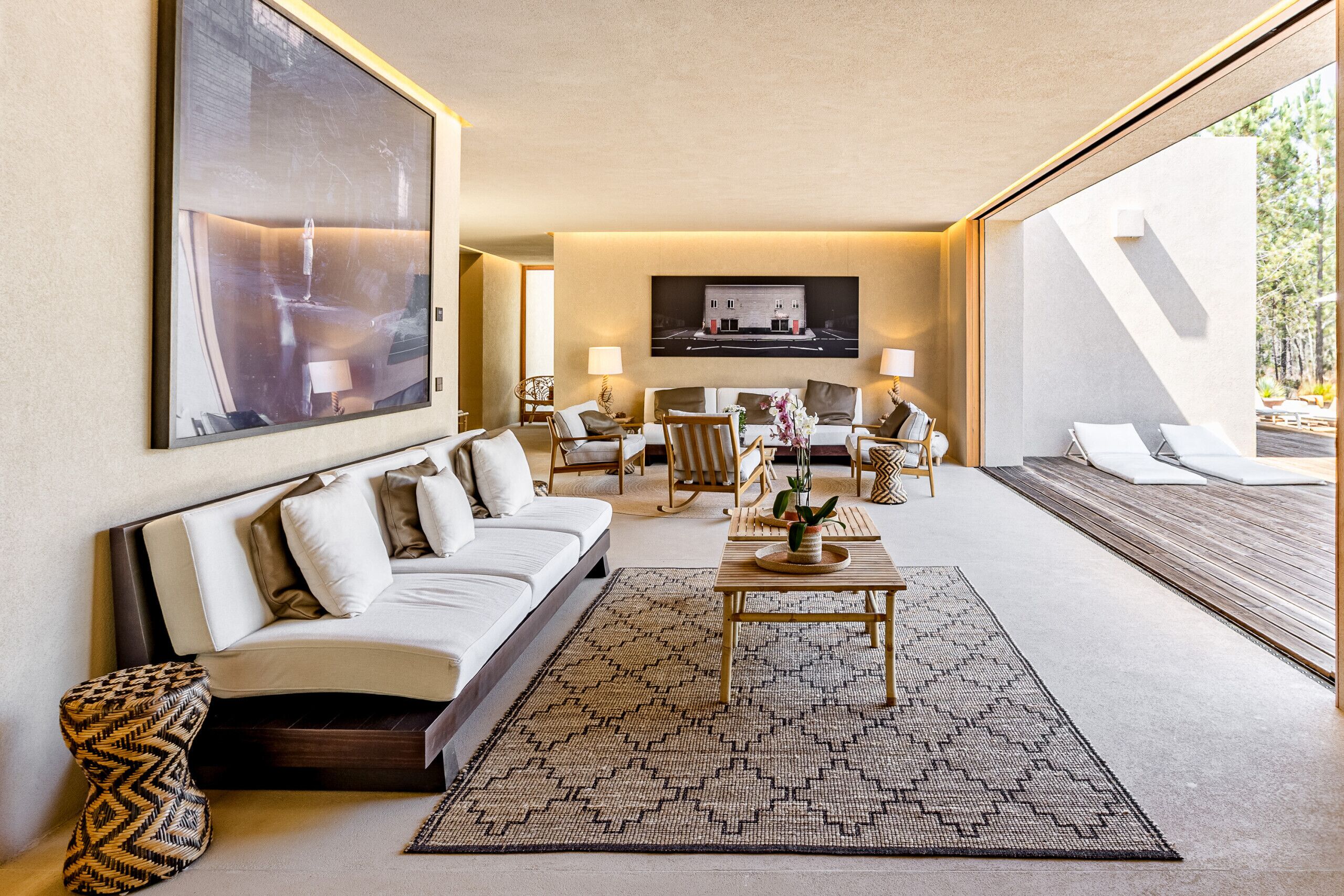 A modern hotel room with a large bed, a sofa, a coffee table, and a seating area with chairs and a small table. There is a large window providing a view of the outdoors.
