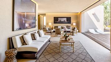 A modern hotel room with a large bed, a sofa, a coffee table, and a seating area with chairs and a small table. There is a large window providing a view of the outdoors.