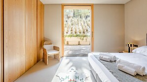 A bedroom with a wooden wardrobe, a wicker chair, a bed with white linens, and a view of a garden through a glass door. - Melides Premium Villa With Pool by Homing (Grandola)