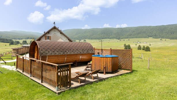 A rustic wooden cabin with a hot tub and deck furniture, surrounded by a fenced yard and green fields.