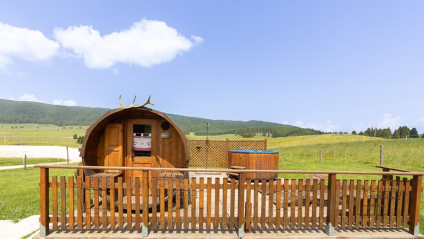 Een houten hut met een houten terras en een omheind stuk grond, omgeven door een grasachtig landschap en heuvels.