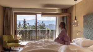 A bedroom with a large bed, a view of mountains through a sliding door, a green chair, and a hanging light fixture. - Hotel Baumwirt (Castelrotto)