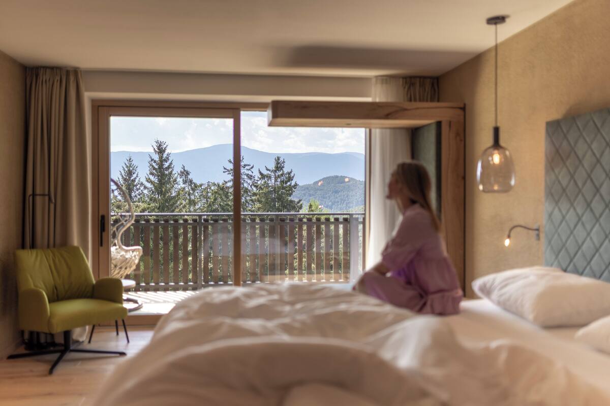 A bedroom with a large bed, a view of mountains through a sliding door, a green chair, and a hanging light fixture.