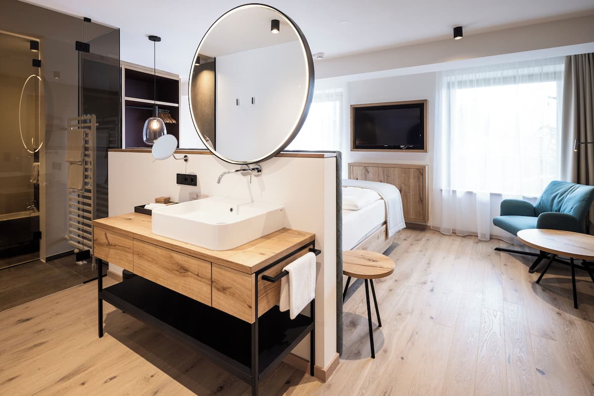 A modern hotel room with a wooden vanity, a large mirror, a bathtub, a TV, a sofa, and a small round table.