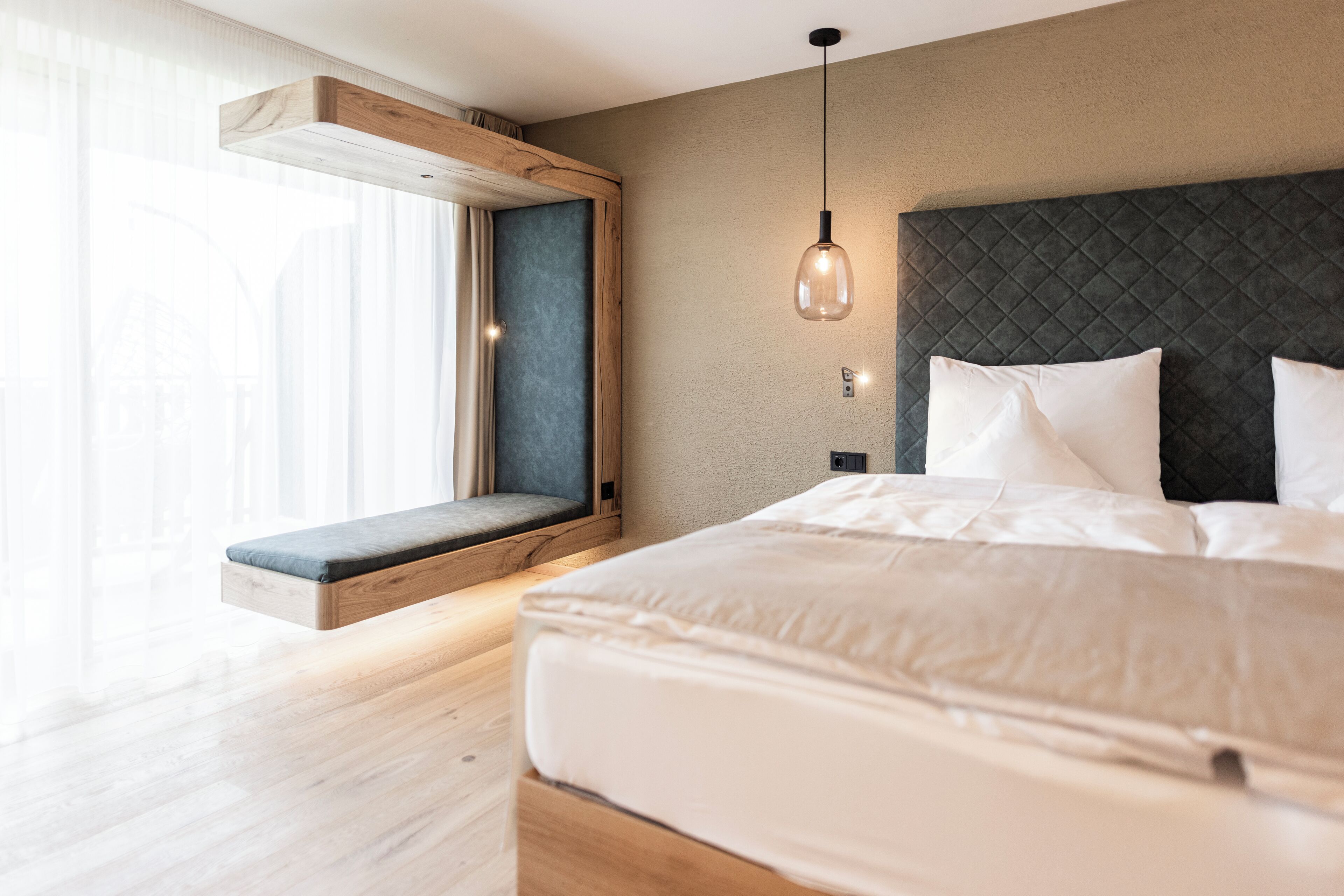 A modern bedroom with a bed, a bench, a hanging light fixture, and a window with curtains.