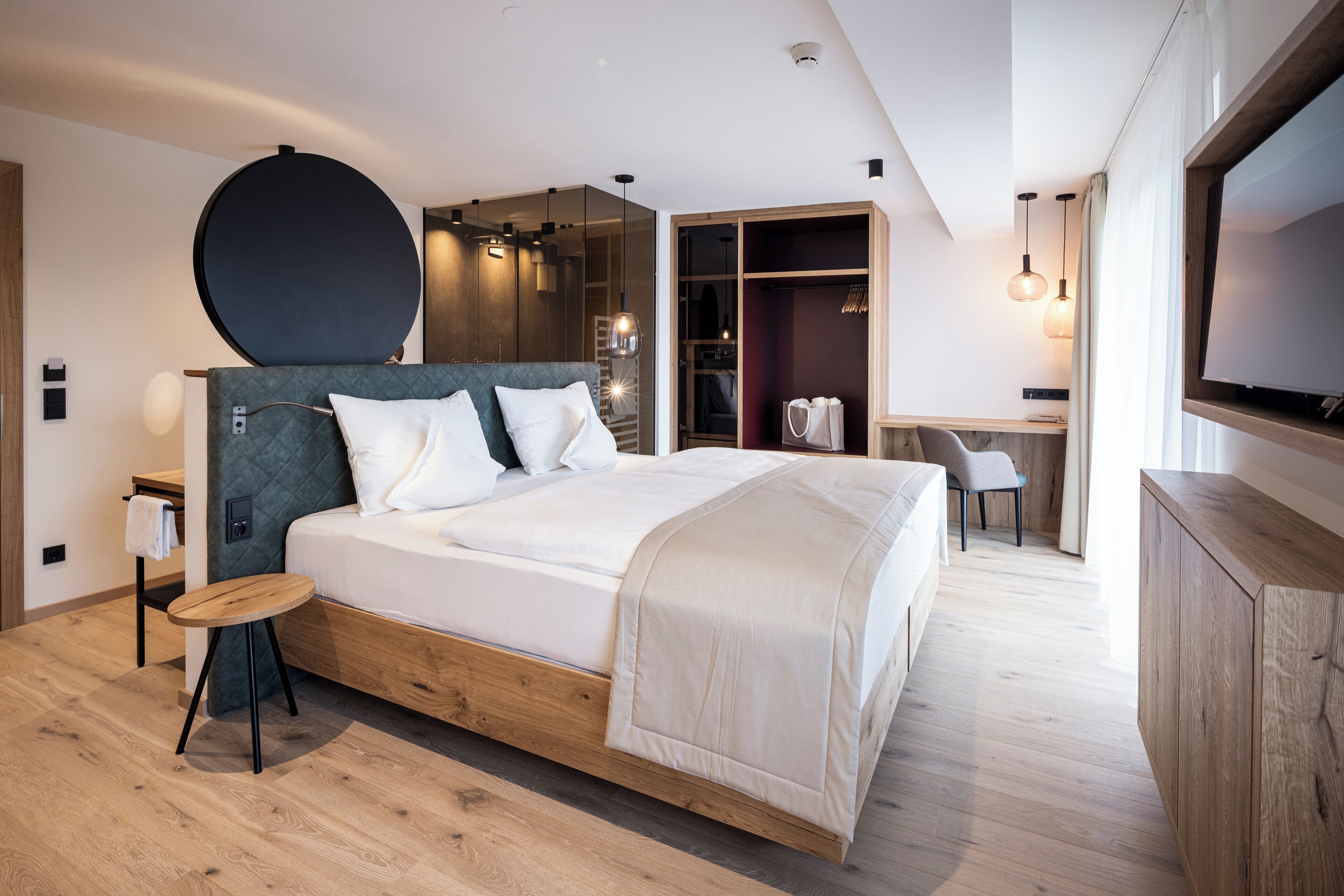 A modern hotel room with a large bed, a wooden headboard, a round mirror, a desk with a chair, and a wardrobe.