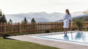 A person standing by a swimming pool with a wooden fence and mountains in the background. - Hotel Baumwirt (Castelrotto)