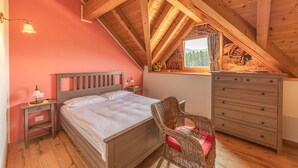 Une chambre avec un plafond en bois, un lit, une commode, une chaise et une fenêtre donnant sur des arbres.