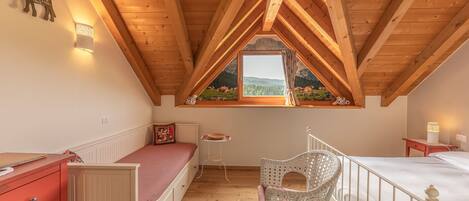 A bedroom with a sloped wooden ceiling, a window with a mountain view, a bed, a chair, and a small table.