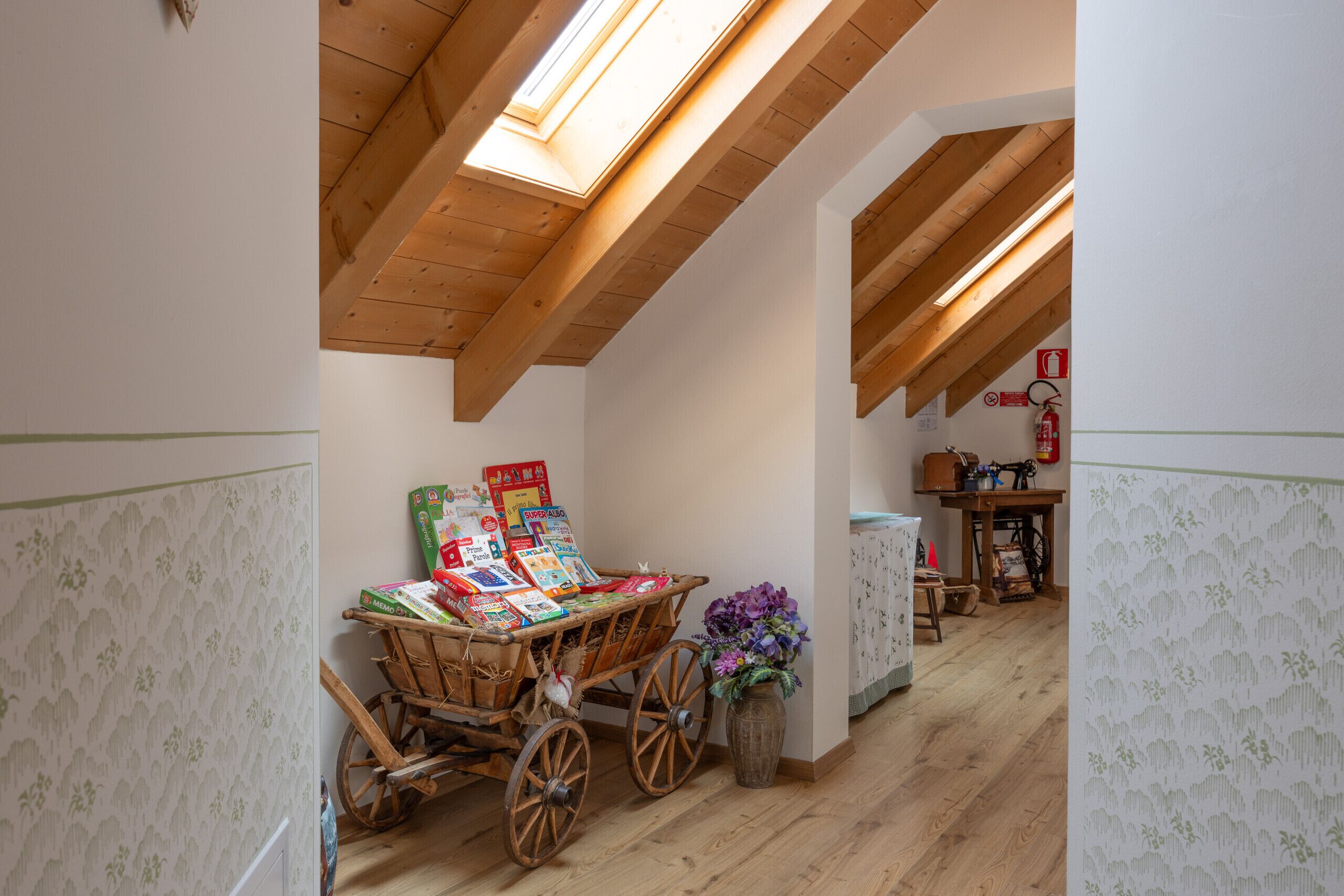 A cozy attic room with wooden beams, a wooden cart with magazines, a vase with flowers, and a vintage sewing machine.