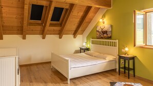 Ein Schlafzimmer mit schrägen Wänden, Holzbalken, einem weißen Bett, einem kleinen Nachttisch mit Lampe und einer grünen Wand mit einem Hirschbild.
