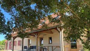 Un espace extérieur aménagé, abrité et doté d’une table et de chaises, d’une structure en forme de pergola, et d’un bâtiment avec un toit recouvert de tuiles rouges en arrière-plan.