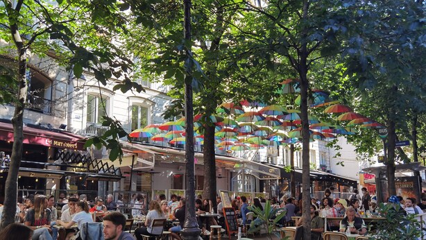 Un café al aire libre con mesas y sillas redondas, rodeado de árboles y edificios.