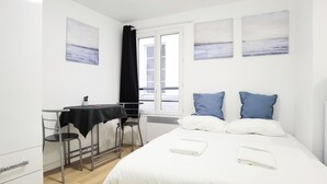 Un dormitorio con cama, dos almohadas, una mesita de noche, una silla, una ventana con cortinas y obras de arte en las paredes.