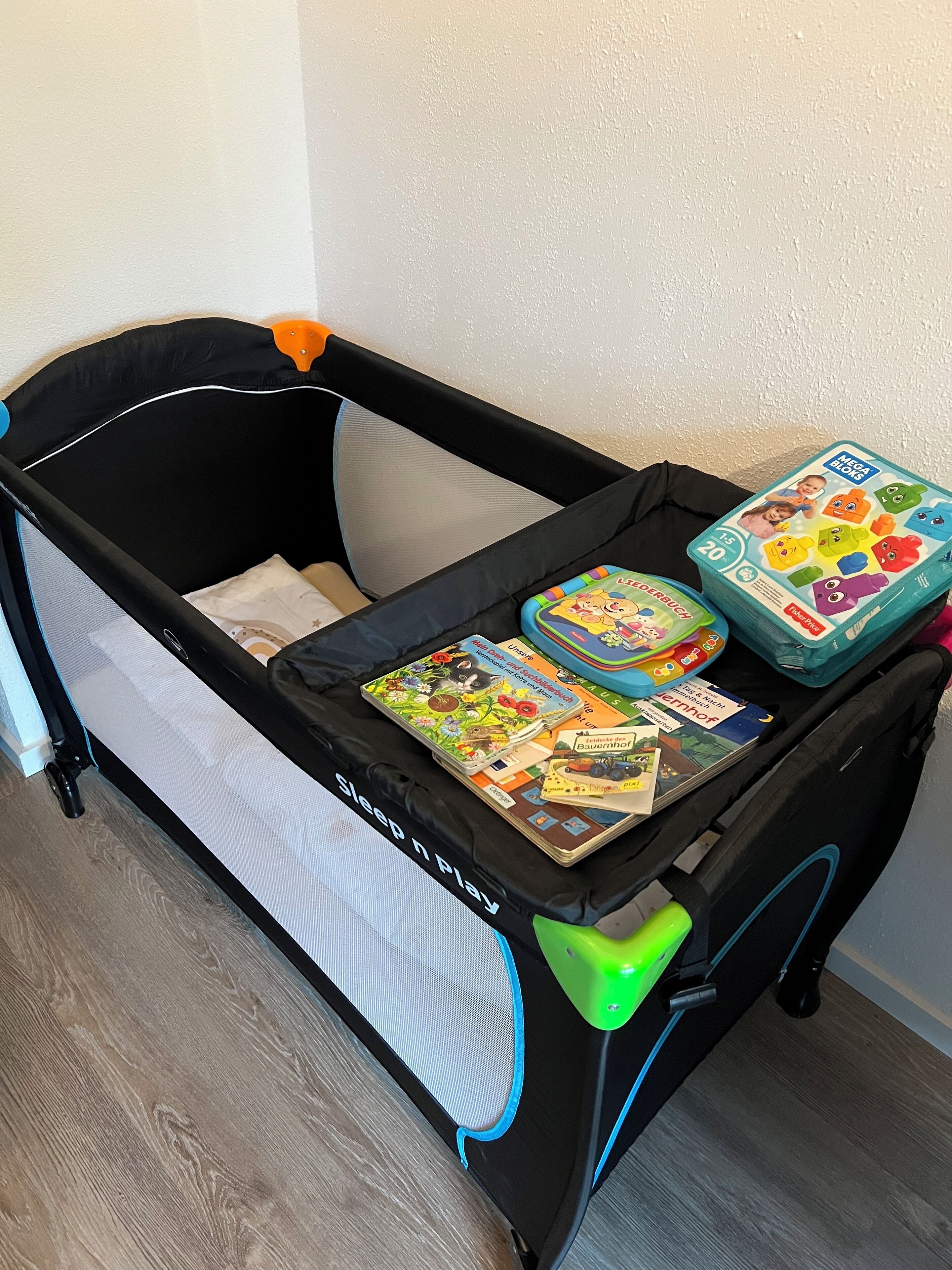A black baby playpen with a sleeping and play area, a green footrest, and a toy box with colorful blocks.