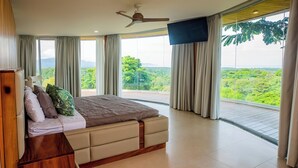 A bedroom with a large bed, a ceiling fan, a TV mounted on the wall, and a balcony with a view of greenery.