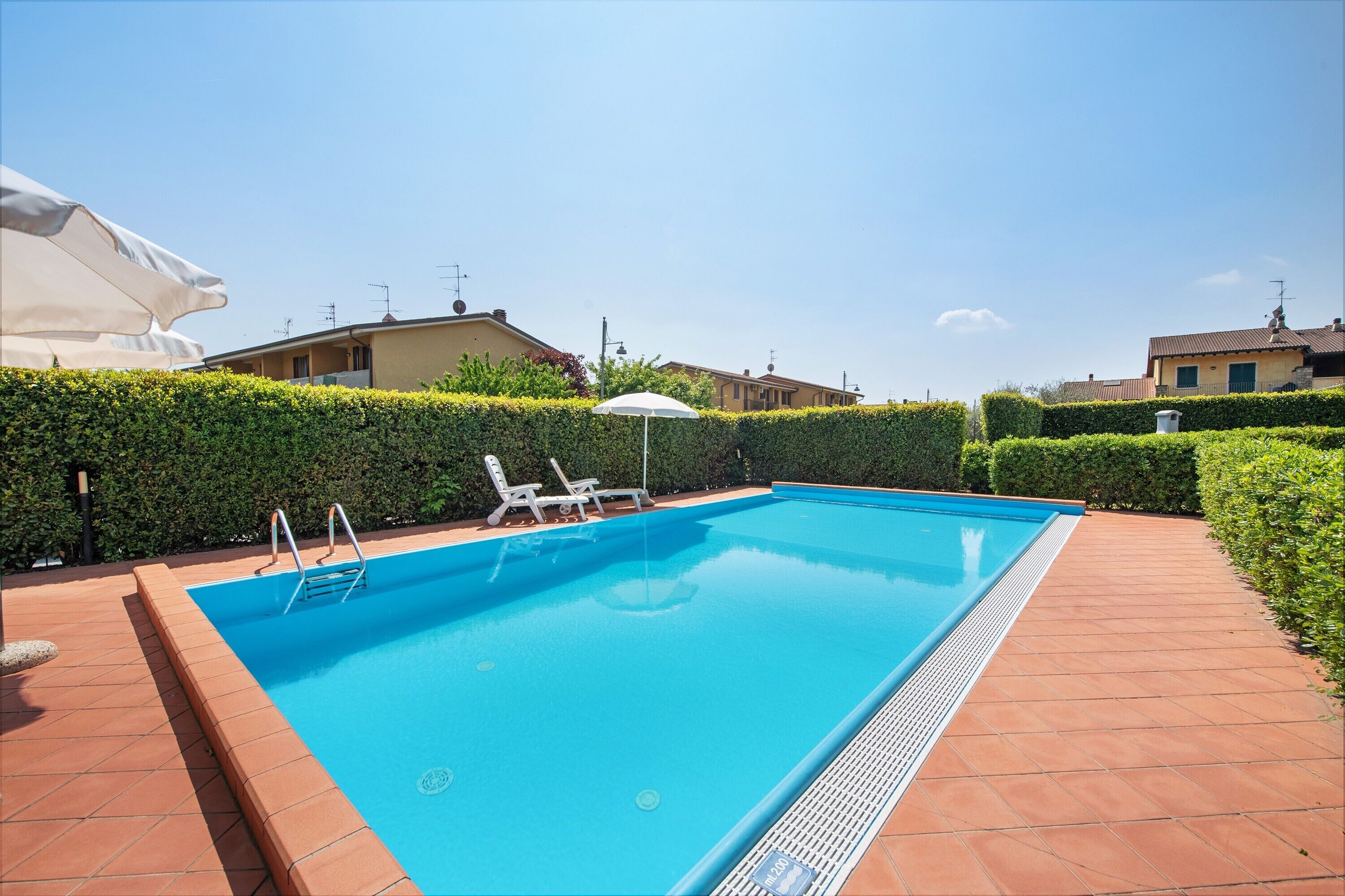 Una piscina rectangular amb vora de rajola, envoltada per un bord de setetes, i al costat d'un edifici amb teulada emblanquida.