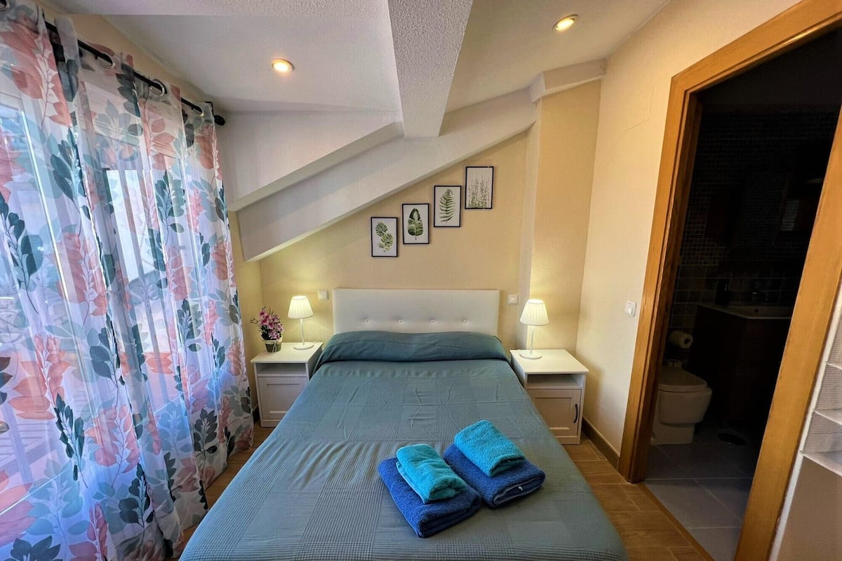 A hotel room with a bed, two bedside tables, a nightstand, a bathroom visible through an open door, and a ceiling with recessed lighting.
