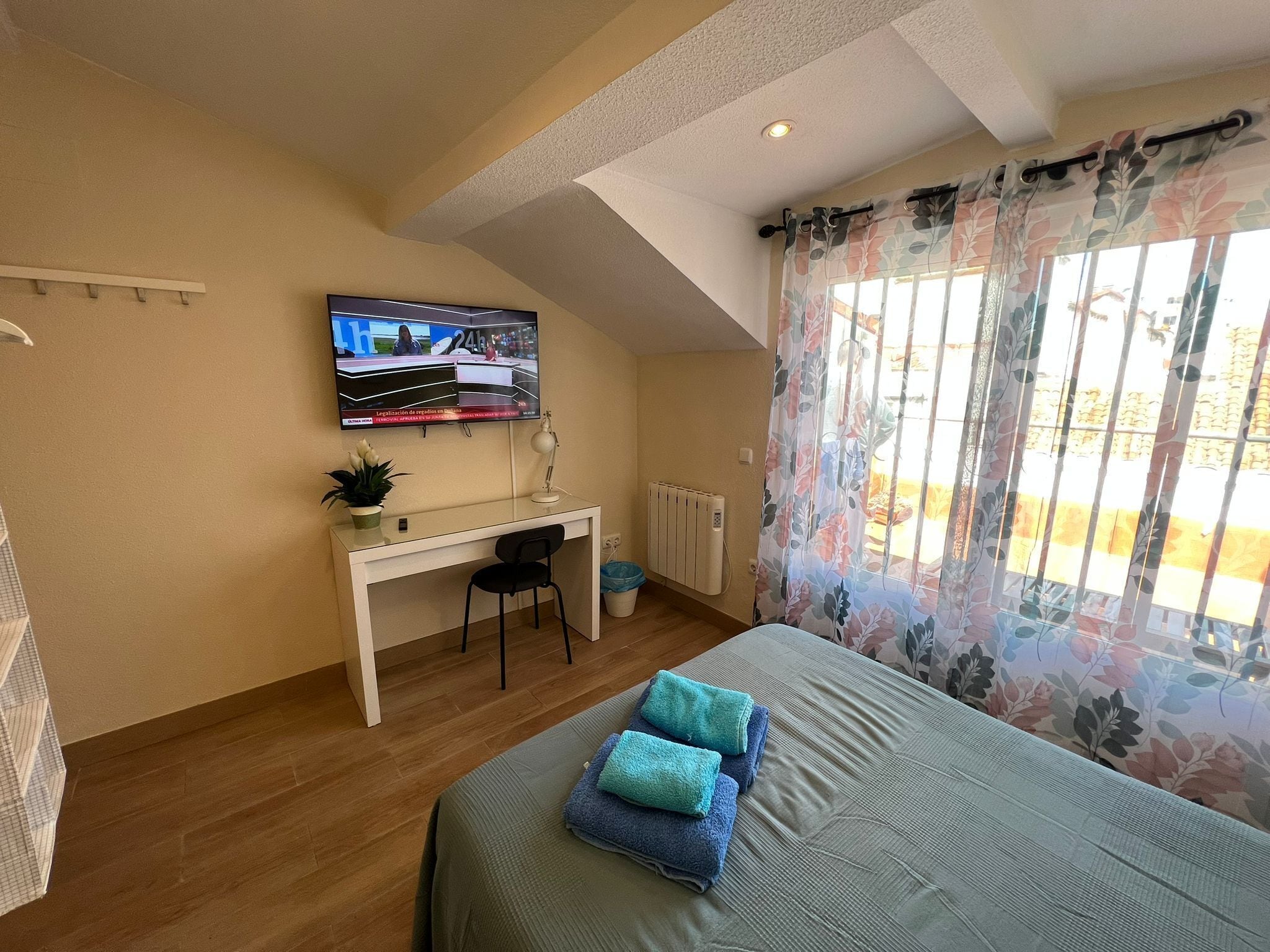 A bedroom with a bed, a desk, a chair, a TV, and a window with curtains.