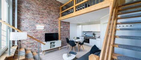 A modern interior with a brick wall, a wooden staircase, a living area with a sofa and a TV, and a kitchen in the background.